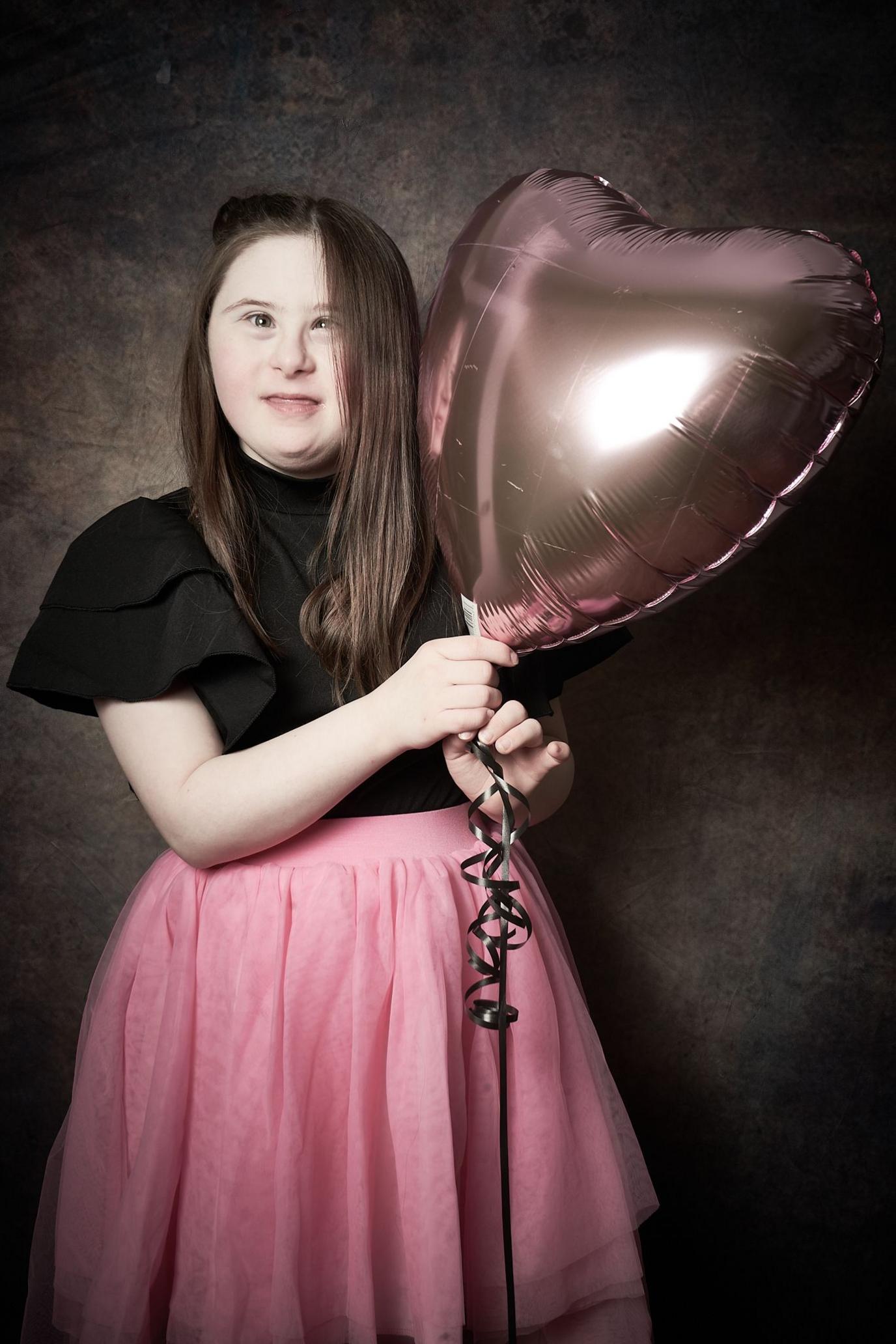 Ava, an older girl with long brown hair wearing a black top and pink skirt holds a large pink heart-shaped balloon.