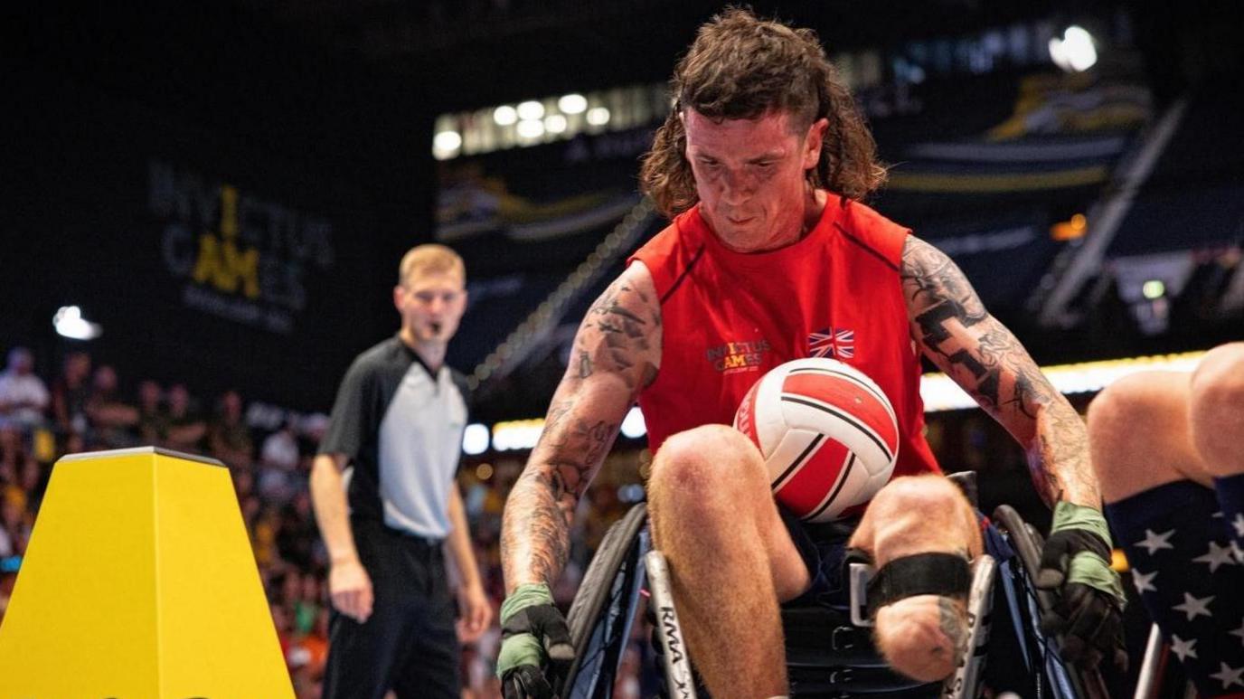 Charlie Holford, a man with a brown mullet hair cut. He wearing a red, sleeveless tank top with sports gloves on and has tattoos on his arms. He is in a wheelchair on a sports court and one of his legs has been amputated below the knee. It is strapped into position on the chair with a black band. On his lap is a white and red ball. The background is blurry, but there is a crowd in the stands and a male referee with a whistle in his mouth.