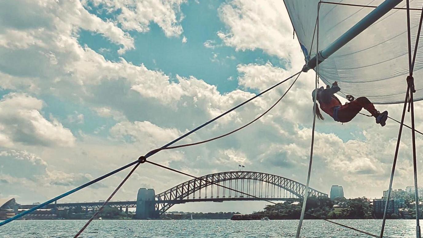 Meg Niblett climbing the rigging of a boat, of which only part of a sail and some ropes are in view on a cloudy but sunny day with a view of Sydney Harbour Bridge in the background