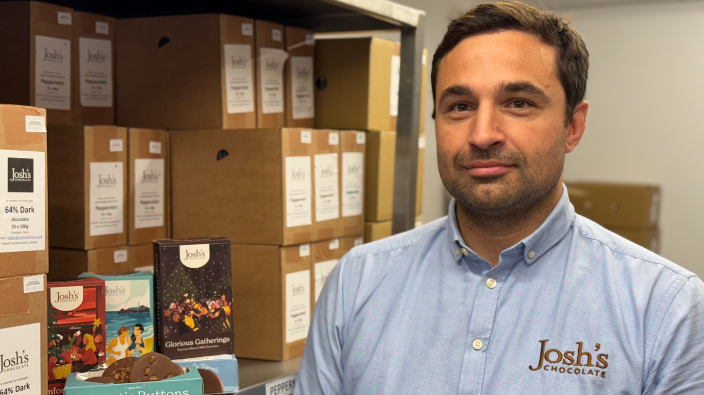 Josh Parker, who is wearing a blue shirt, is stood in his stock room. On one side of him there are boxes of chocolate products with some open and on display on shelves.