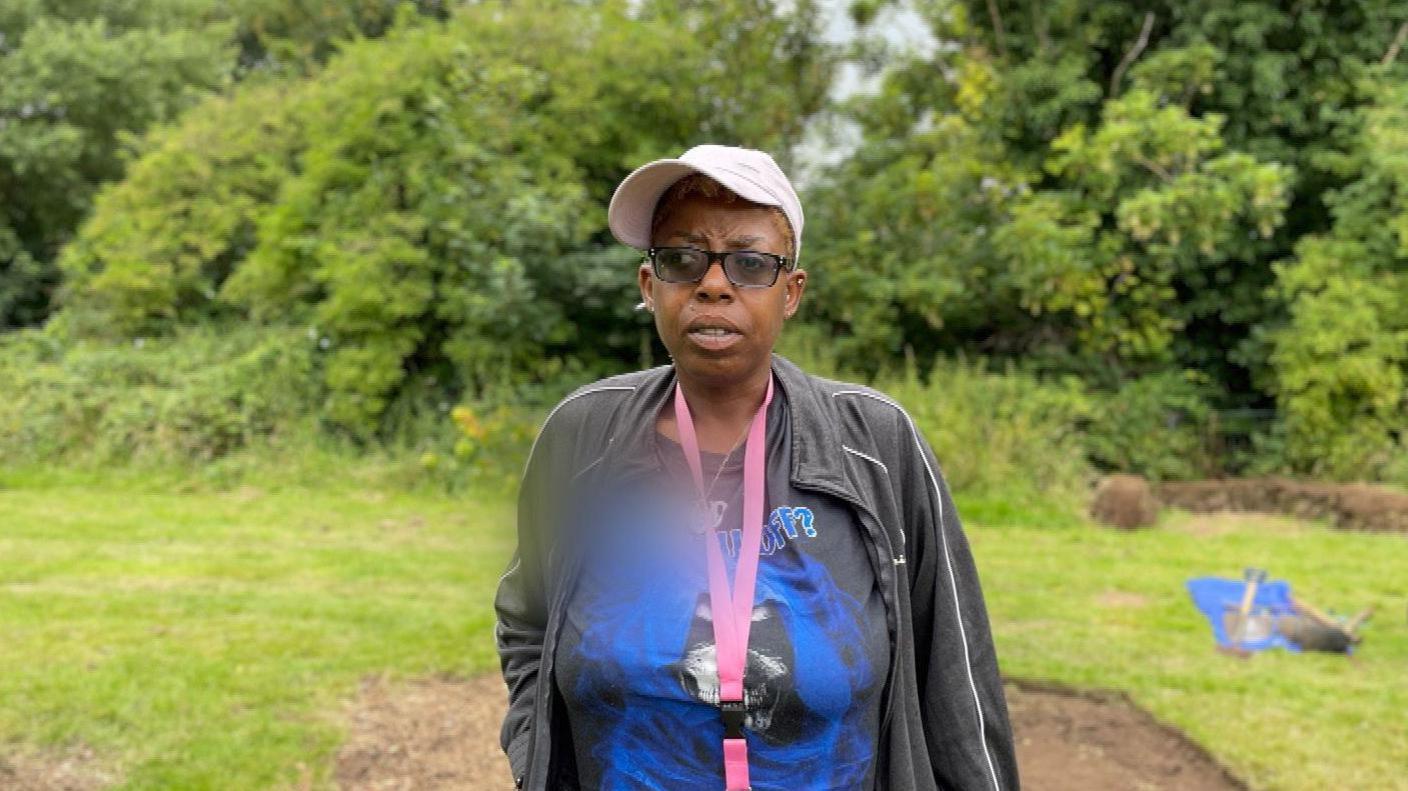 Chrissie Elliot standing in front of trees at one of the sites in Balsall Heath. She is wearing a black jacket, white cap, sunglasses and a pink lanyard.