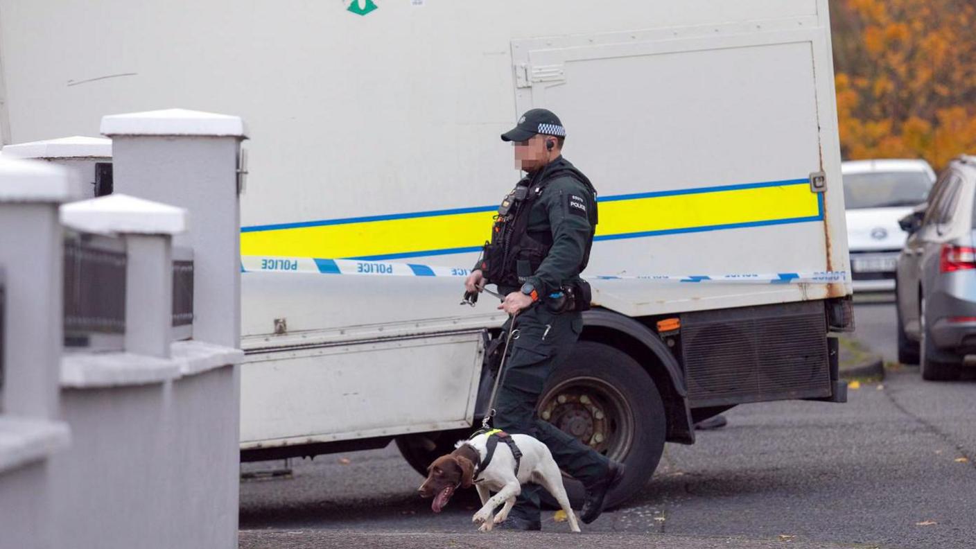 A police officer walking beside blue and white police tape with a dog on a lead. He is wearing a bottle green uniform with a black vest. There is a white van with a yellow stripe up the side beside the tape.
