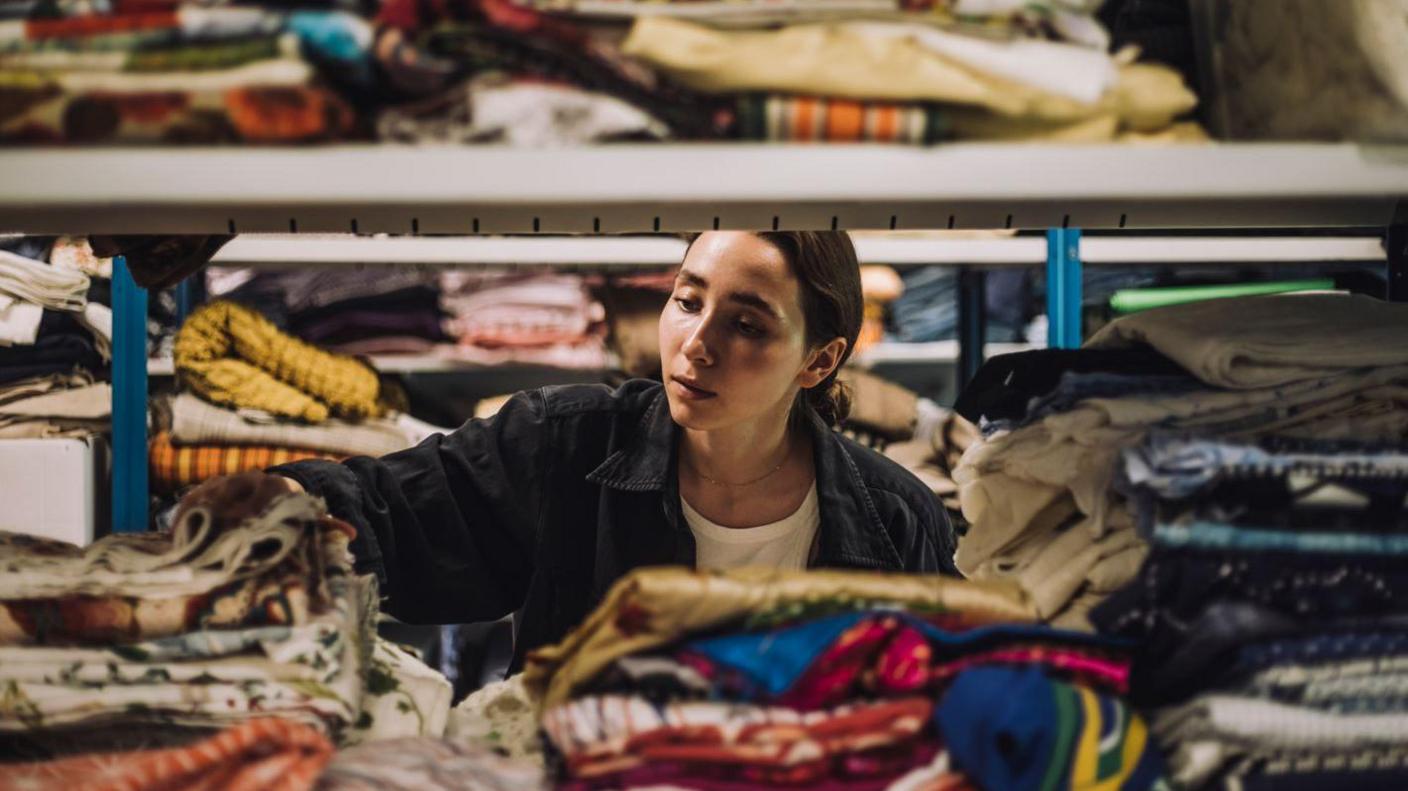 A stock picture of a woman wearing a denim jacket looking through piles of clothes and fabric. She can be seen through a metal shelving unit. There are piles of folded fabric on the shelves in front and behind her.