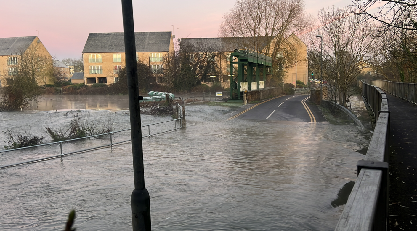 Flood water as taken over a road which can no longer be seen. Towards the back of the image the road re-appears as it goes over a bridge. To the right of the image is a sign for Little Paxton. The sunrise is pink in the sky and there is steam coming off the water in the background.