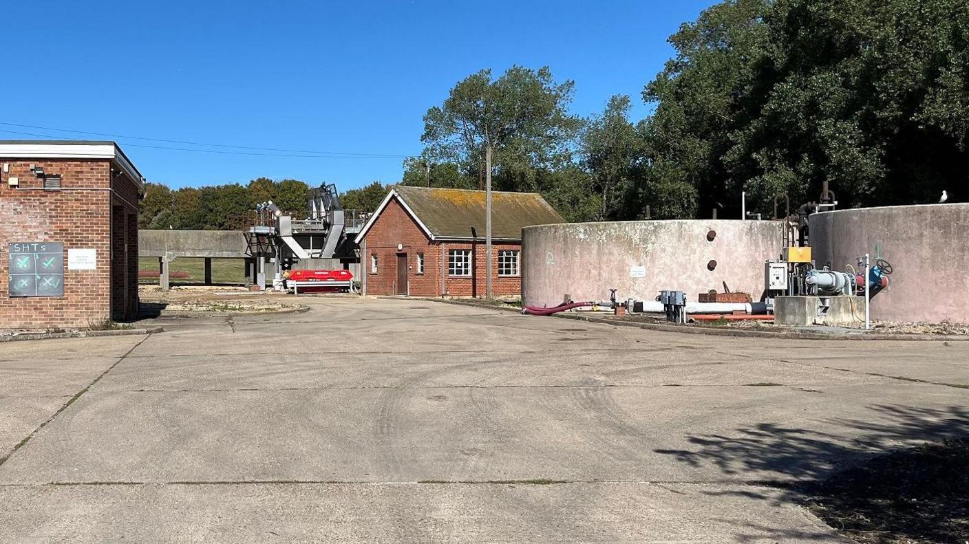 Water works. On the left is a square brick building. On the right are large circular ponds for processing sewage.