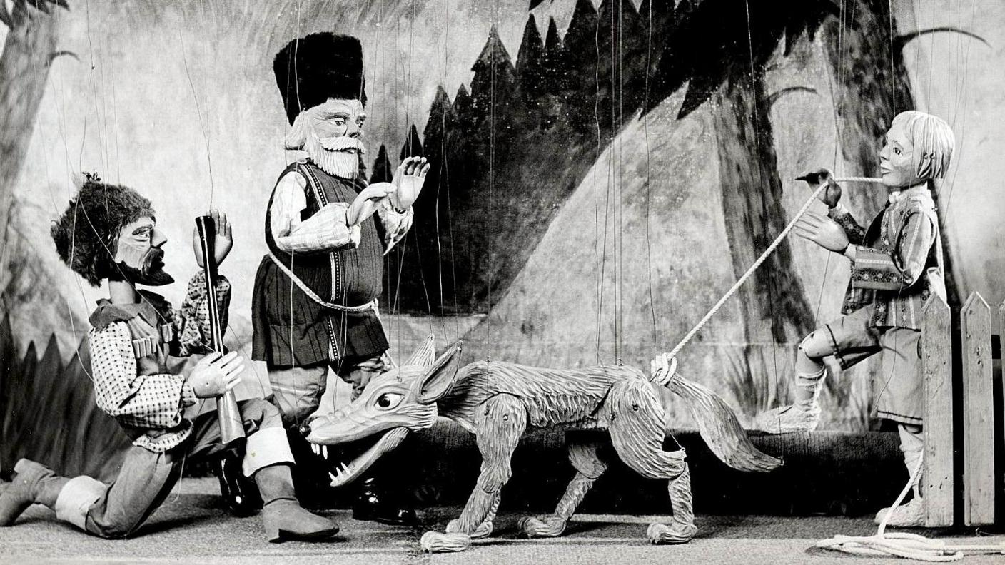 A black and white photograph of puppets on a stage, suspended by string. There are two men, one with a long-barrelled gun, a boy and a wolf.