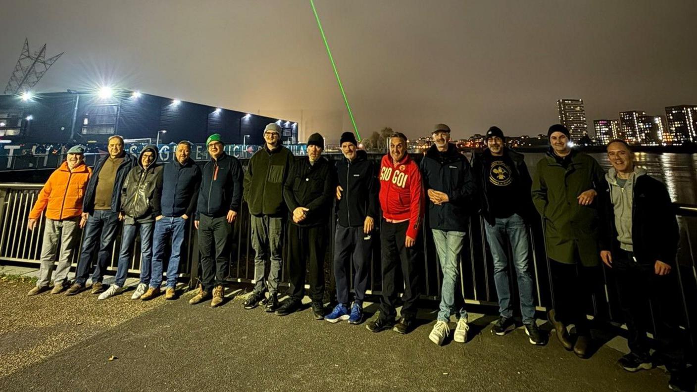 A group of men on a bridge looking at the camera. They wear warm clothing and the sky is dark.