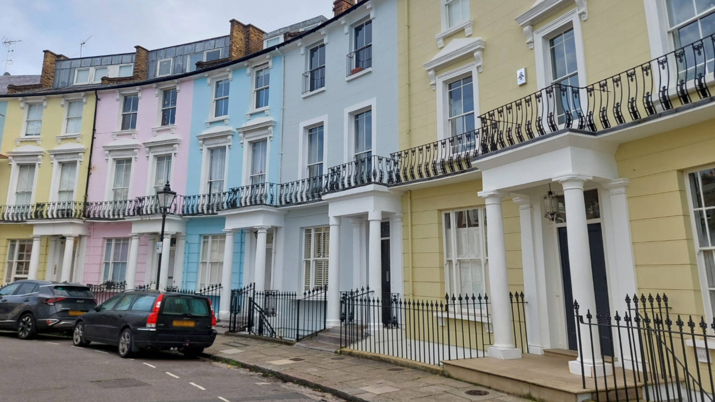 A row of colourful townhouses in Chalcot Crescent, Primrose Hill, London. The houses are each four or five floors tall and are yellow, pink, blue and grey. There are two black cars parked outside.