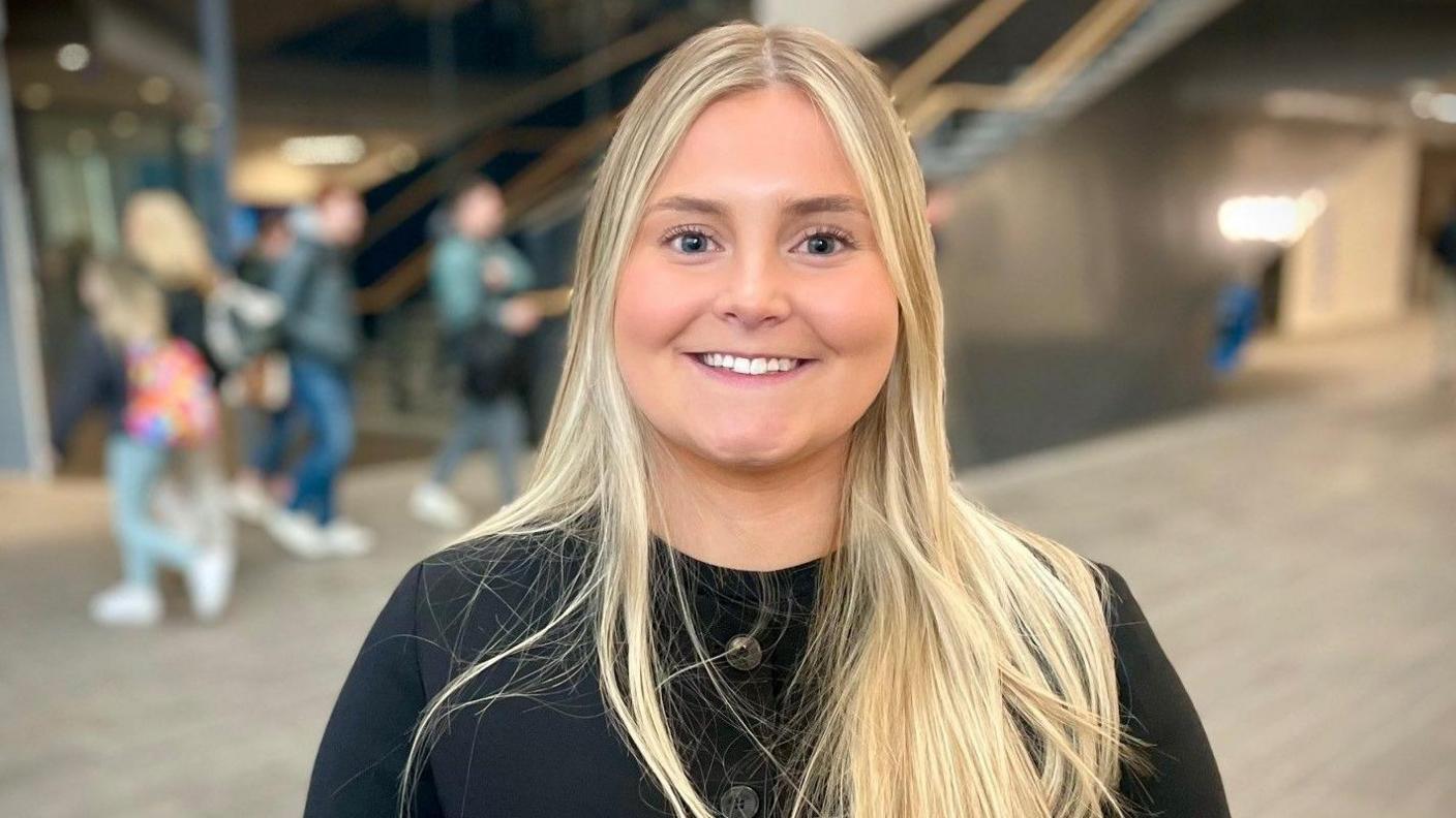 Anna Dukelow - a young woman with long blonde hair smiles at the camera. She is standing in the middle of the university campus building where students are walking in the background but it is blurred. She is wearing a black buttoned-up cardigan.