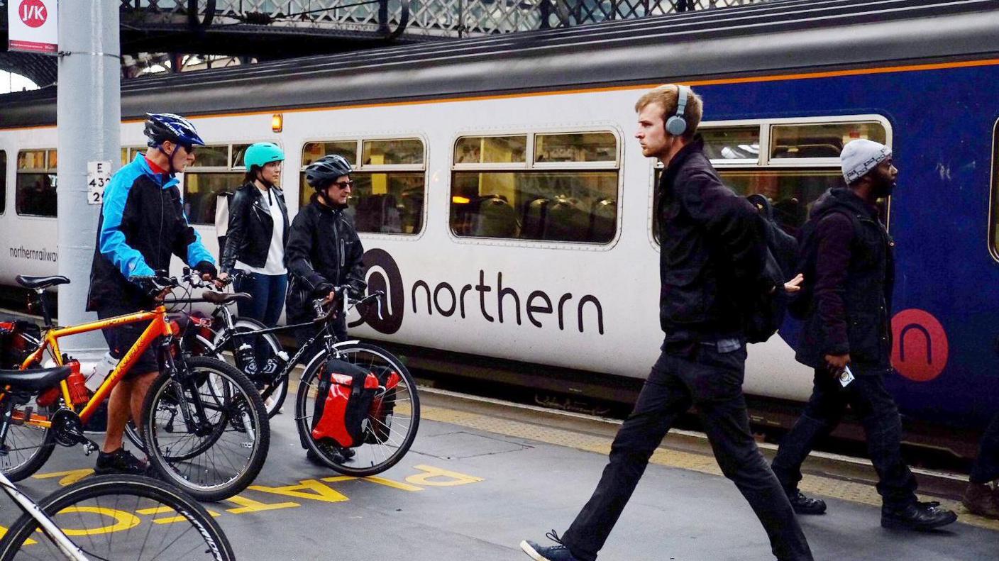 A train at a platform. Several passengers in cycle clothing push bikes towards the train whilst a man in a black jacket and headphones strides away in the opposite direction.