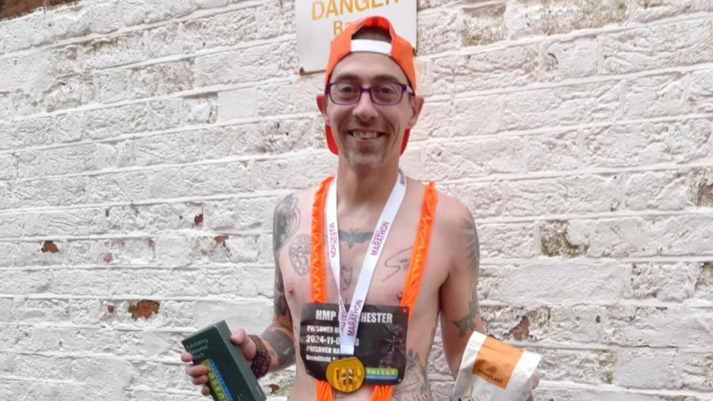 A smiling David Andrewartha wearing an orange mankini and back-to-front orange baseball cap. He is wearing glasses and has a number of small tattoos on his chest and arms and an orange medal around his neck. He is posing in front of a whitewashed brick wall and holding snacks in his hand..