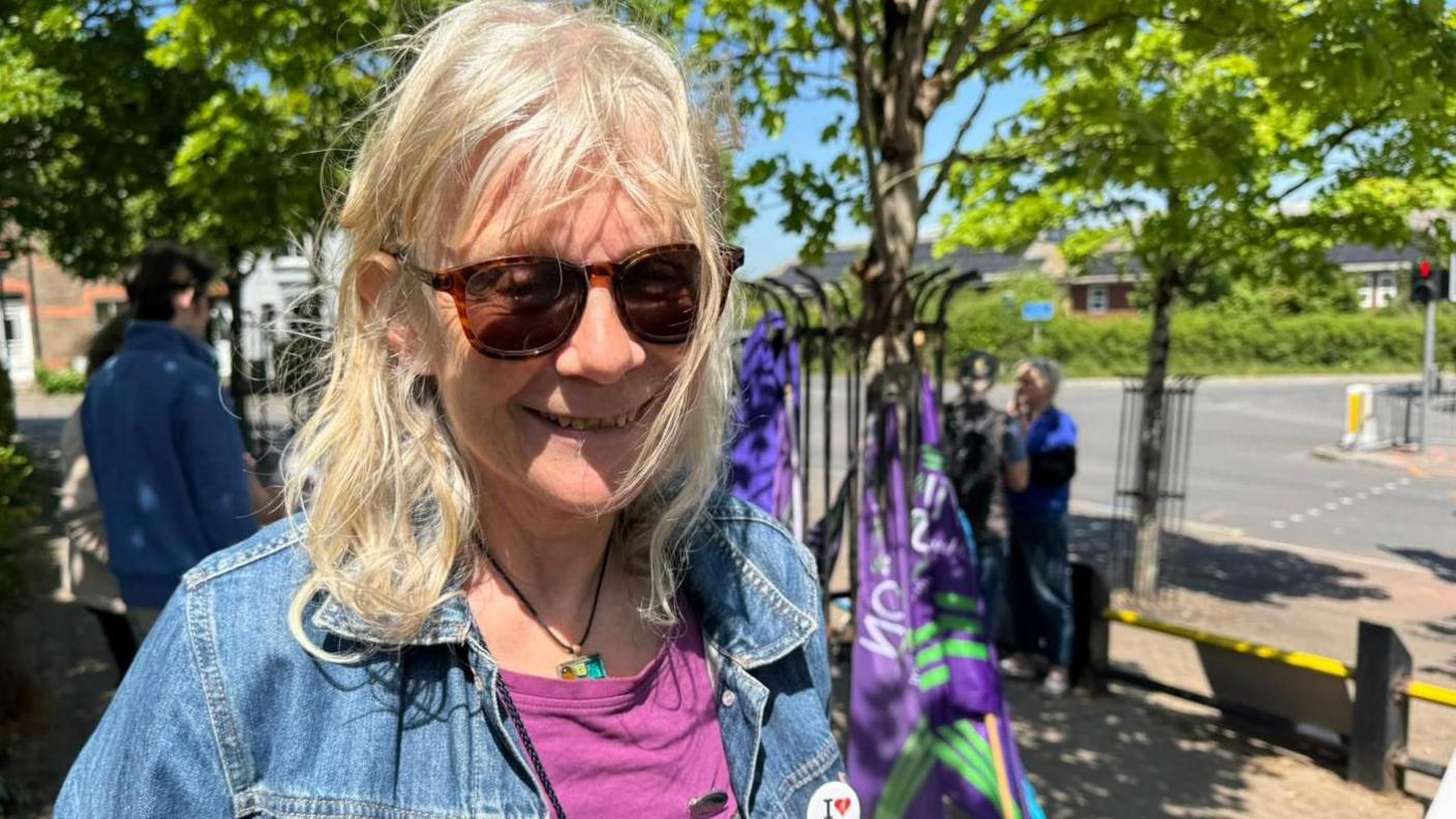 A smiling Lynne Hubbard at the protest in Dorchester. It's a sunny day. She is wearing sunglasses and has shoulder length blonde hair. She is wearing a denim jacket and a cerise t-shirt.
