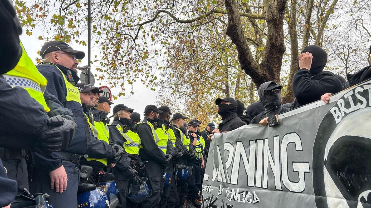 Police line in front of counter-protesters at Saturday's demonstration. The protesters are all wearing black with covered faces and sunglasses. Officers are wearing high vis yellow jackets on black uniforms.