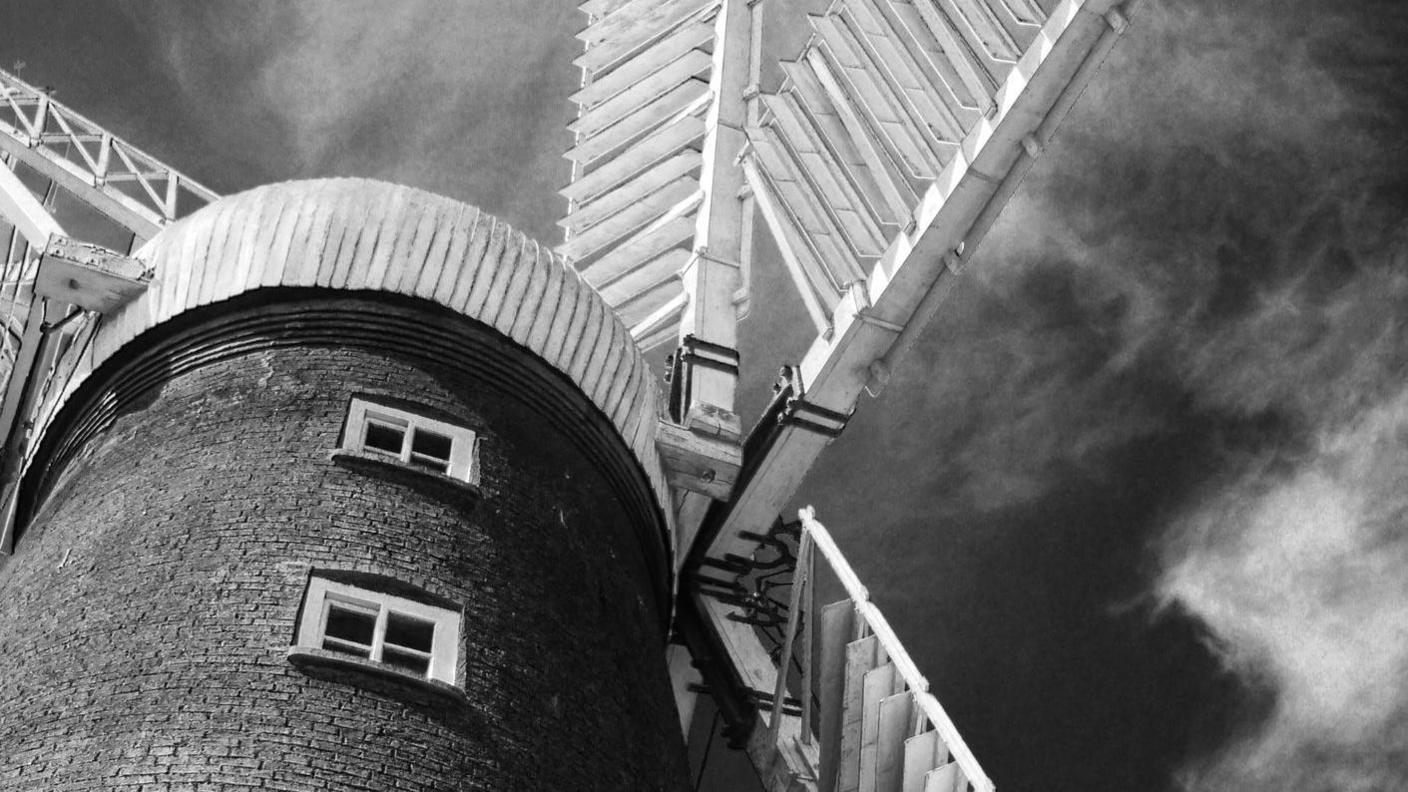 A black and white photograph of Alford Windmill with it's signature white sails and white cap. Bunches of white clouds are above the mill.