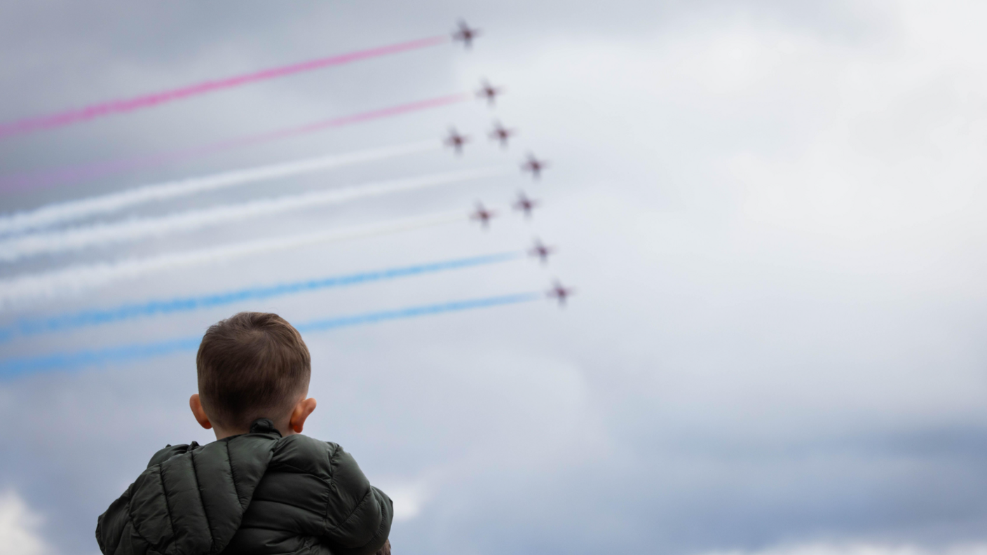 A young boy, seen from behind, looking up at the sky as nine aeroplanes fly through it. Behind them they are leaving red, white and blue vapour trails.