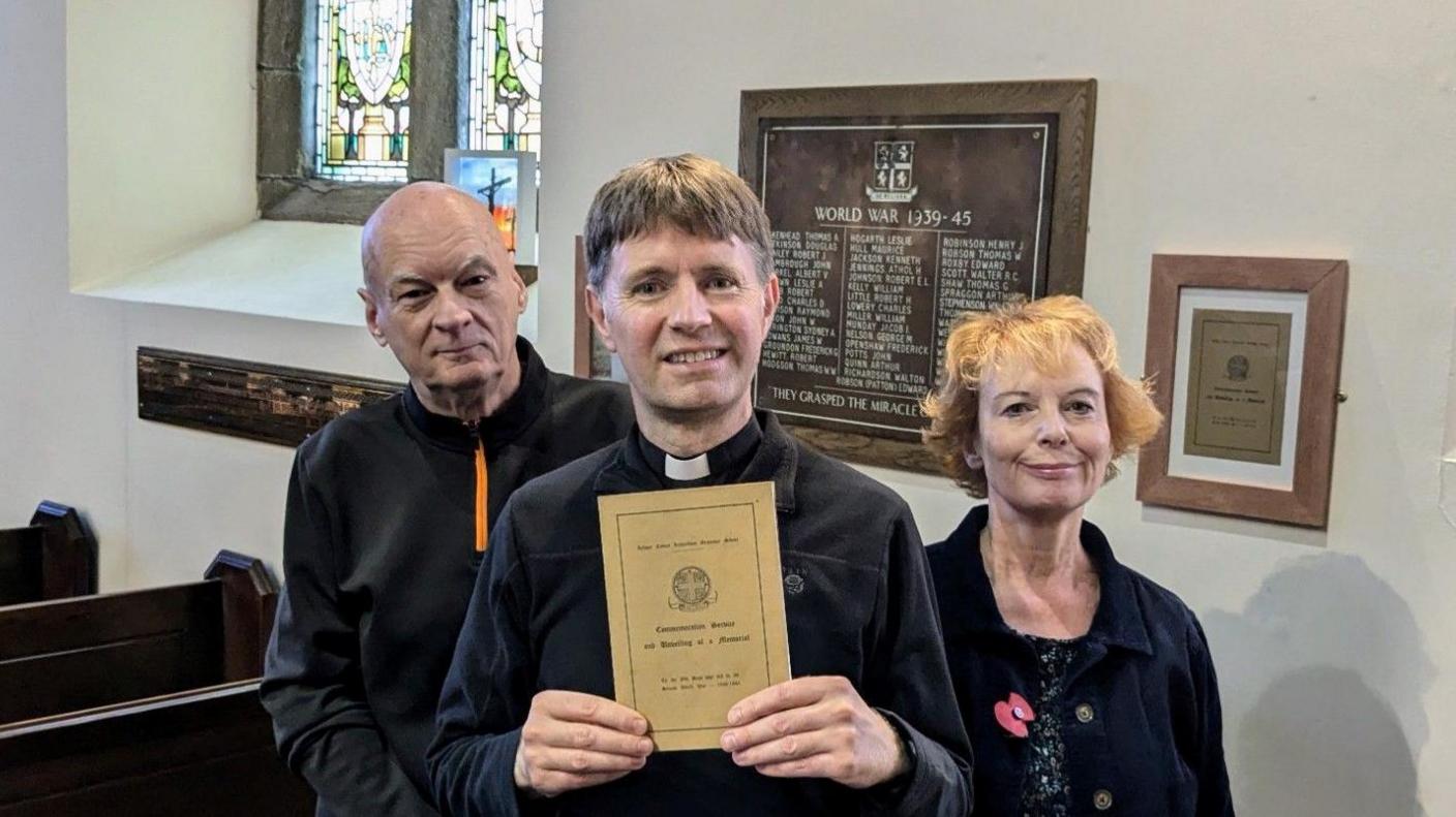 David Chadwick, holding an order service dating back to 1949, stands with two other people in front of a plaque newly installed on the wall of St Paul's, Ryhope 