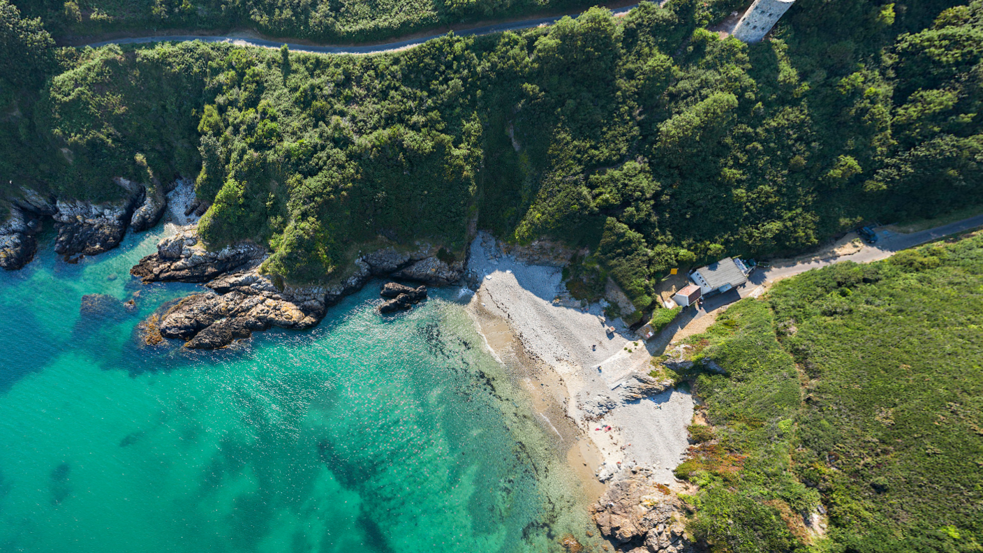 Saint's Bay is a small cove with turquoise waters and a pale sandy beach. There is a path and a stone turret on the top of the cliffs which are lined with shrubs and grass.