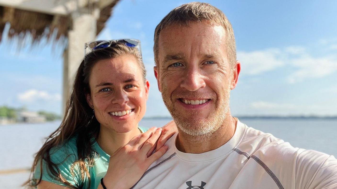 Charlotte Rochenard-Taylor and her husband, Jamie, smiling. They are facing the camera and by the coast. She has long brown hair and sunglasses on her head. He has short fair hair and stubble.