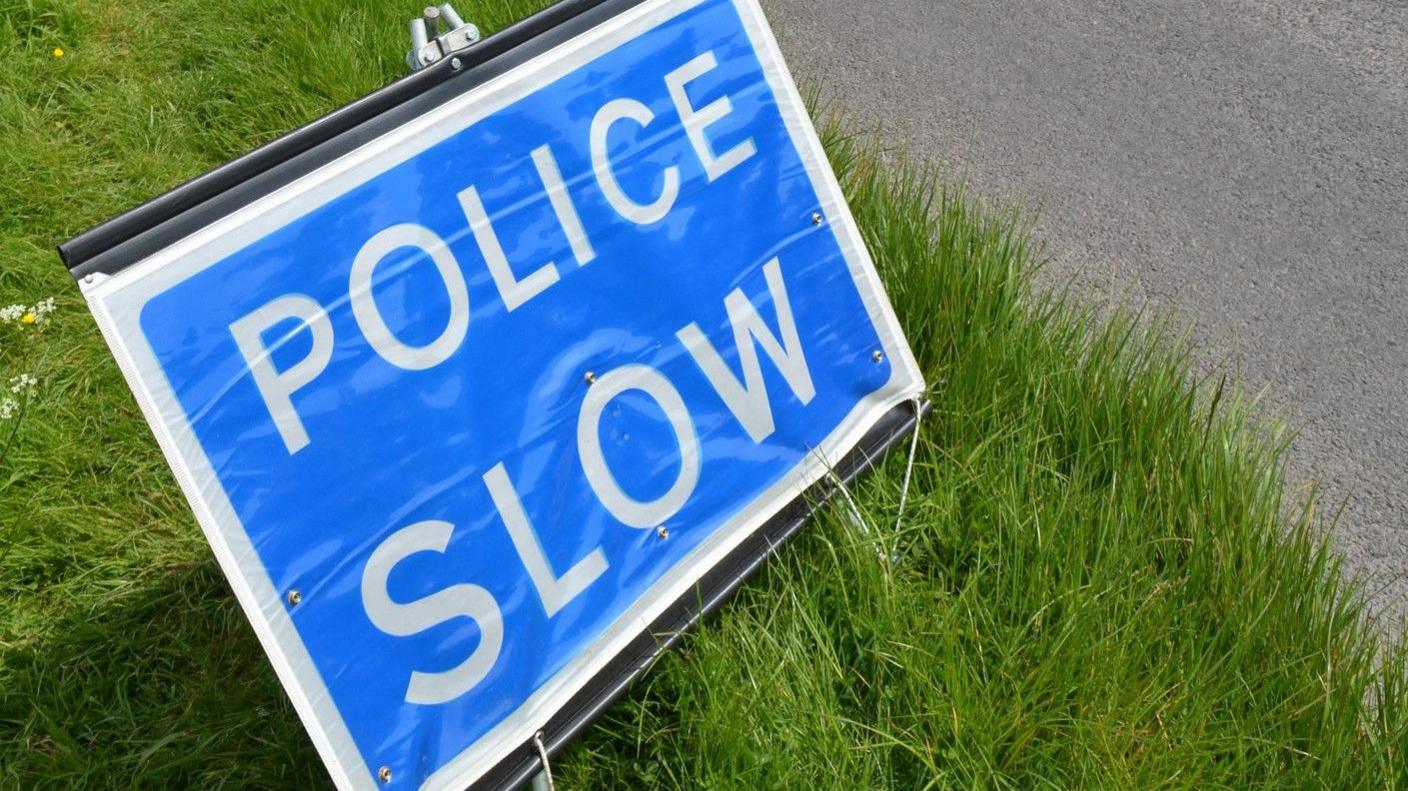 A blue "police slow" sign a grass verge on the side of the road