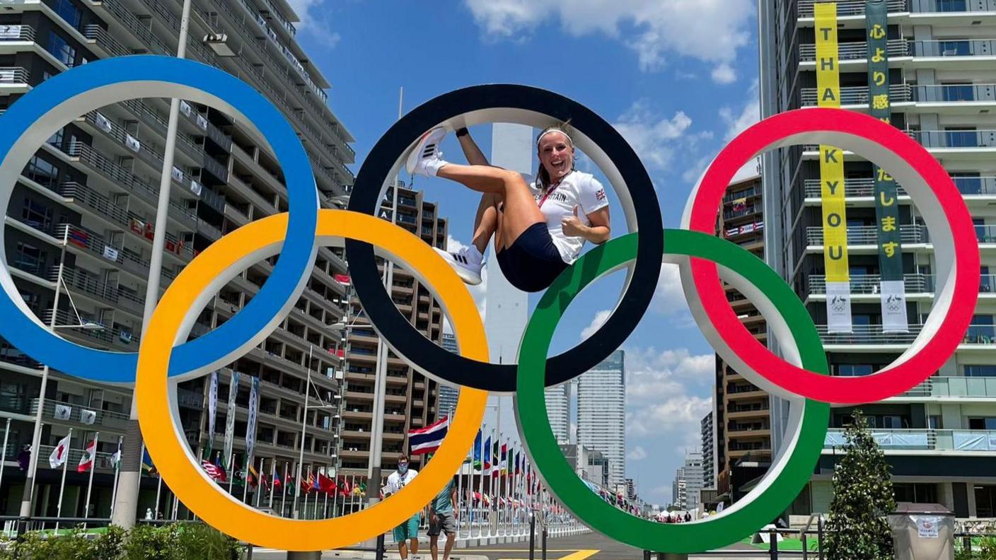 Laura is sat in the middle of the black Olympic ring, the top middle ring, backed by a Tokyo cityscape. Laura is smiling at the camera. 