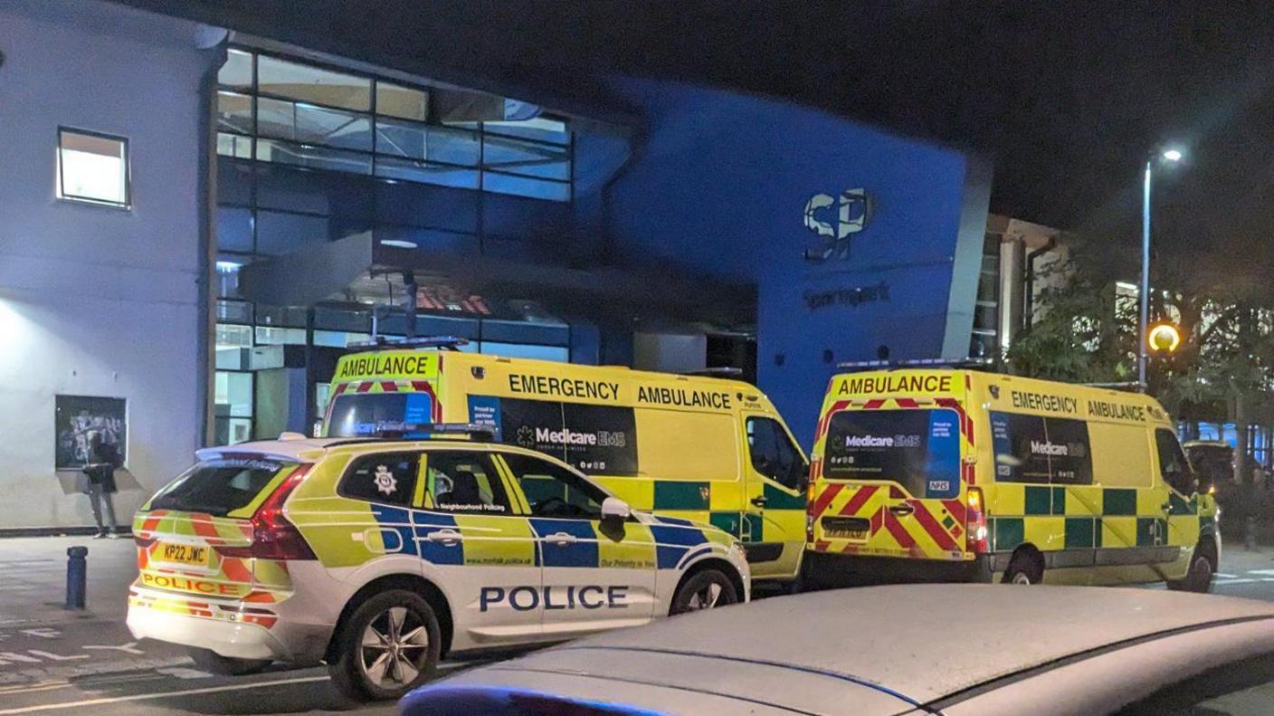 A view of one police car and two ambulances parked outside of the UEA Sportspark. It is nighttime and there is another car parked nearby. The Sportspark building can be seen in the background.