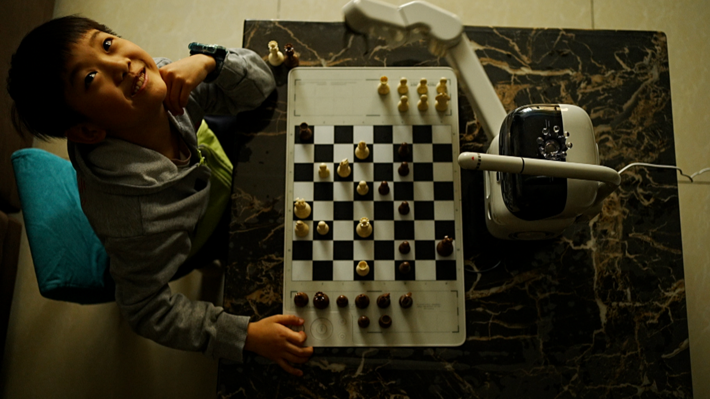 A smiling Timmy looks up at the camera. In front of him on the table is a chess board and a small robot with a white body and a black screen.     