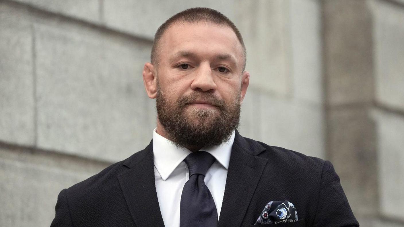 Conor McGregor wearing black suit and tie and white shirt staring at the camera. He has a dark beard and short dark hair.