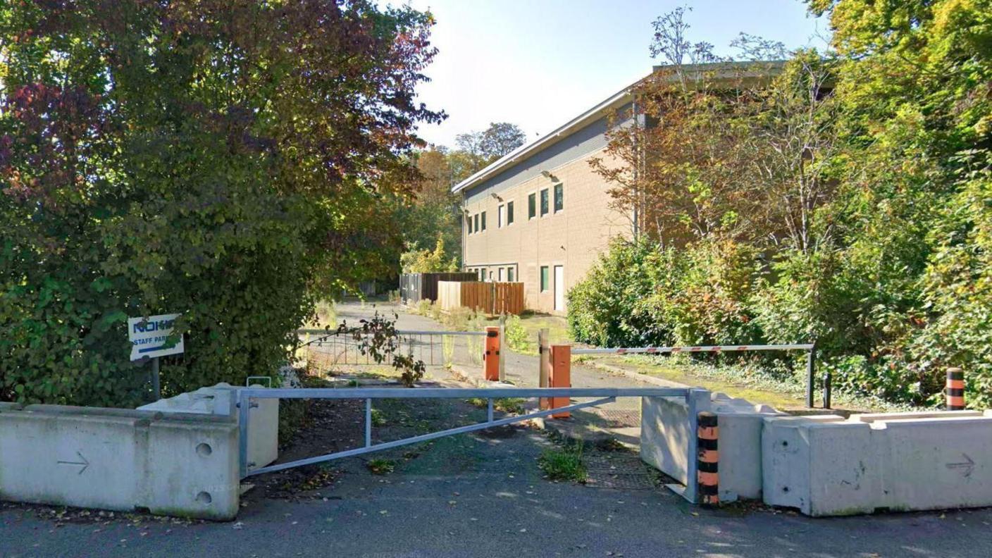 A two-story office building, behind trees and a low wall. There is a car park full of weeds. It is daylight.