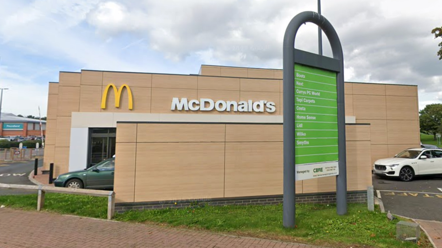 A view of the side of a McDonald's restaurant. There are cars at the drive through, a car park behind, and a green sign showing details of other shops.