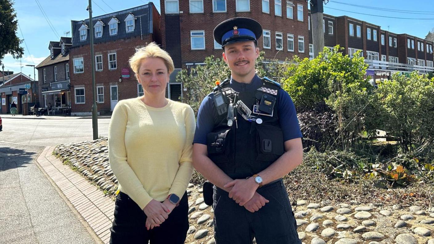 A woman with short blonde hair wearing a pale yellow jumper and black trousers stands next to a man wearing a police community support officer uniform with a body camera and radio attached.