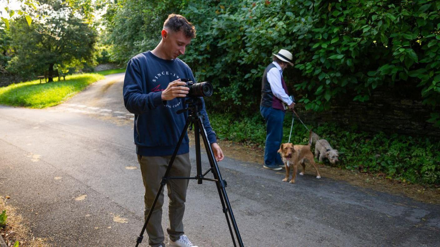 Robin Shuckburgh and Ross Arrowsmith filming on a countryside road. Mr Shuckburgh is looking after his dogs while Mr Arrowsmith is setting up the camera. It's a sunny day.