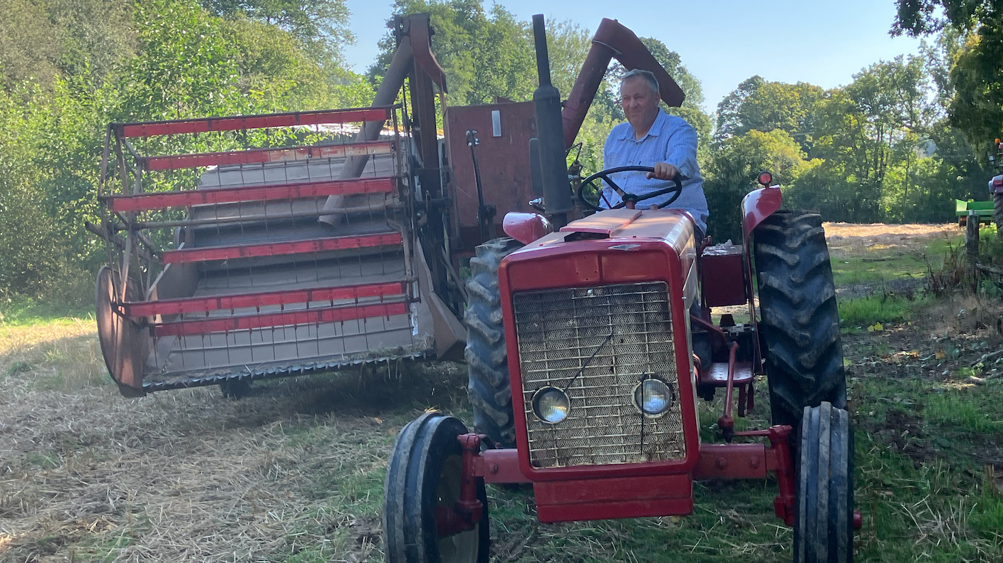 The younger John Hosking sat on a small red tractor towing a McCormick B64 combine harvester in a field.