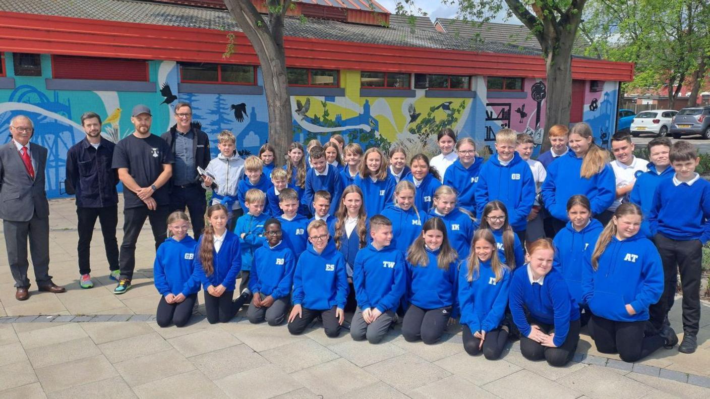 A primary school class all in blue jumpers. Three men stand on the left of the group. Behind them is a colourful mural featuring birds and tress.