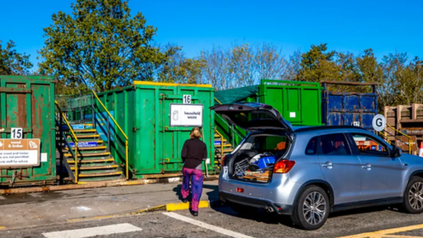 A lady is standing at the boot of the car and looking towards four storage containers that are used for waste at a recycling centre.