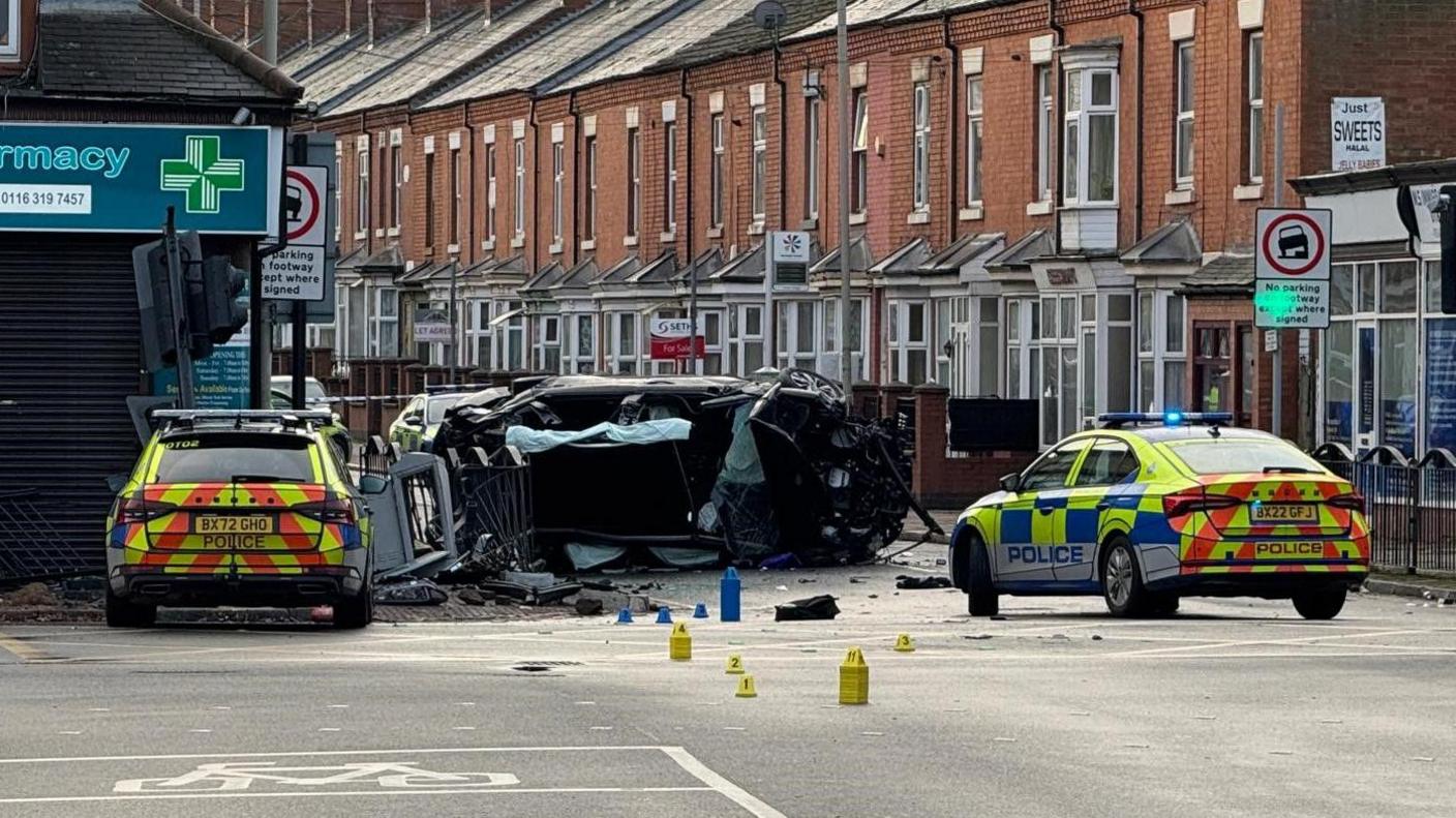 Two police cars at a junction. Two damaged cars, one flipped on its side can be seen, with debris in the road