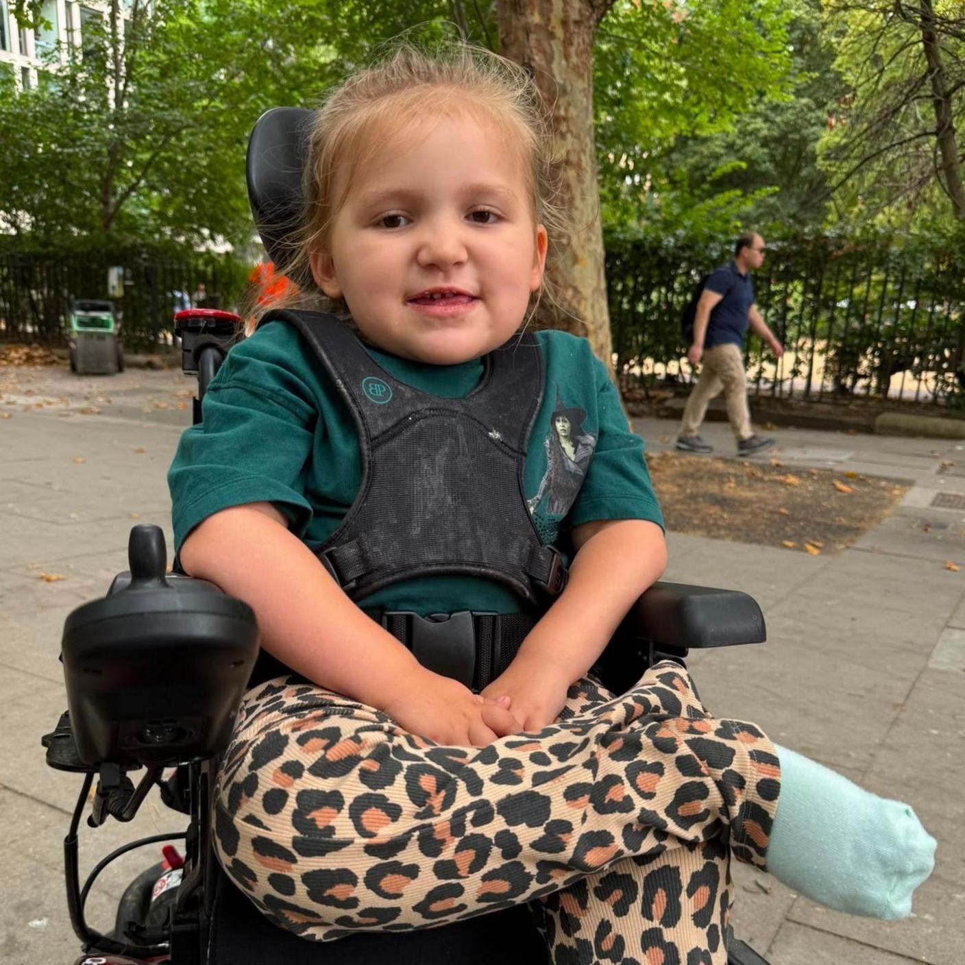 A young blonde girl in a black wheelchair with black straps across her chest. She is wearing a green Wicked t shirt and leopard print trousers. Behind her is a pavement and green-leaved trees.