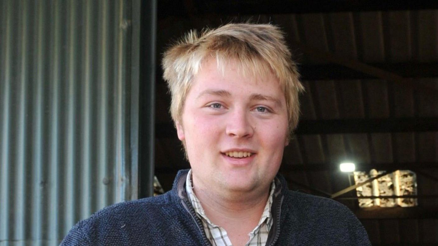 Harry Sukling smiles at a camera. He has blond hair and is wearing a navy jumper over a checked shirt. Behind him is a farm building.