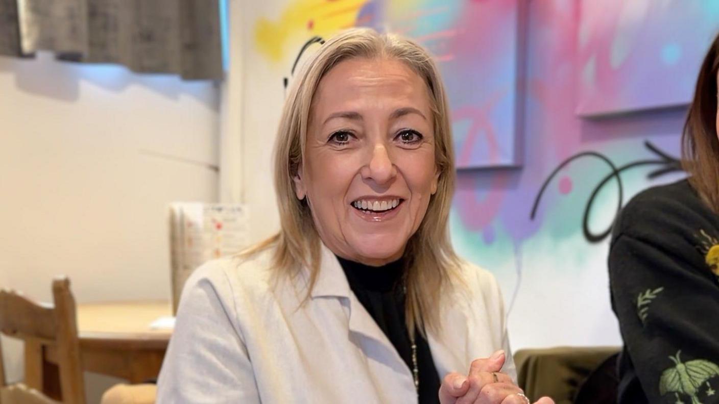 Sallie looks into the lens smiling. She has blonde hair and wears a black turtle neck with a white blazer. She is clasping her hands.