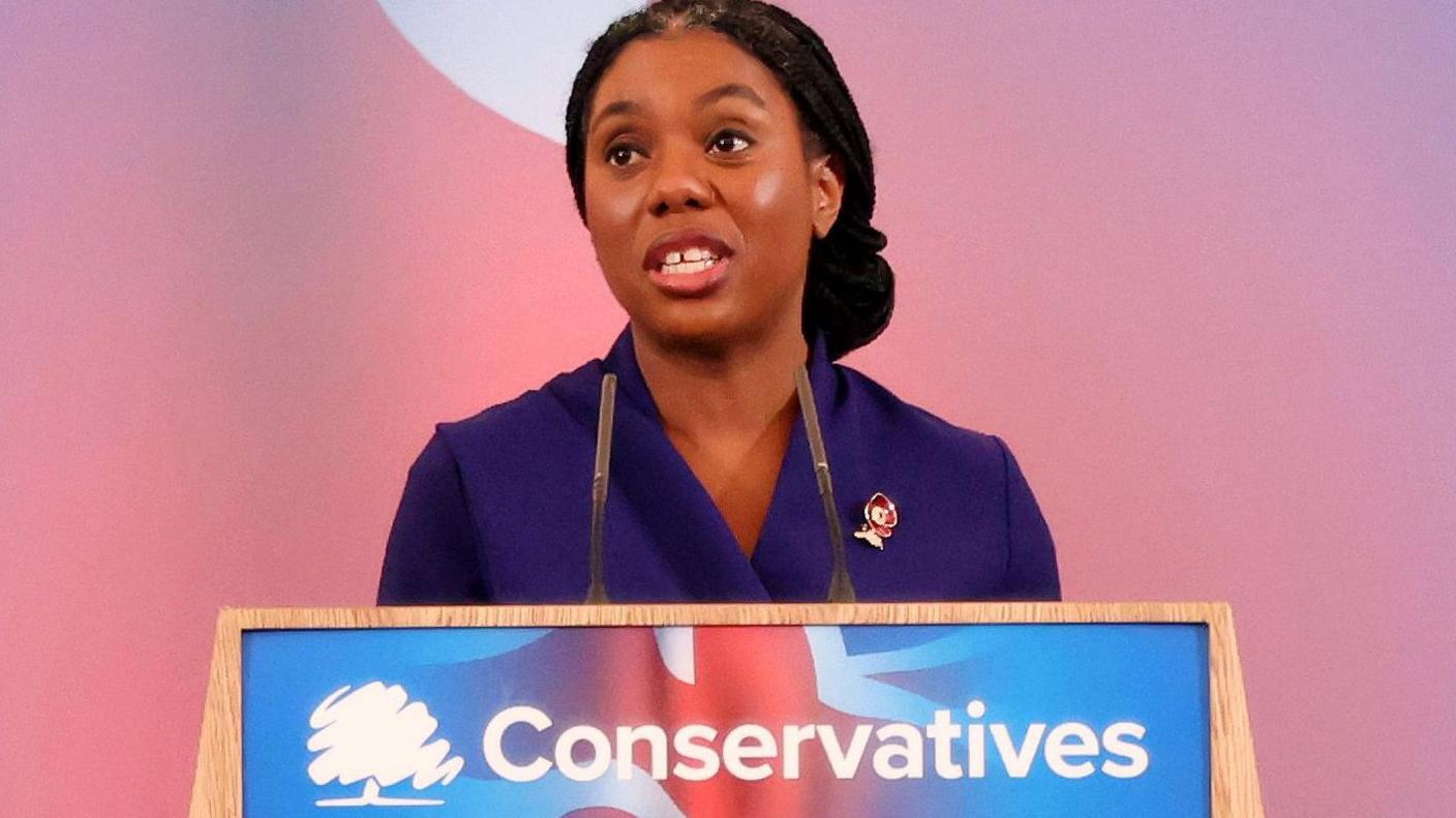 Kemi Badenoch standing in front of a plinth with a Conservatives logo on it. She is wearing a purple dress.