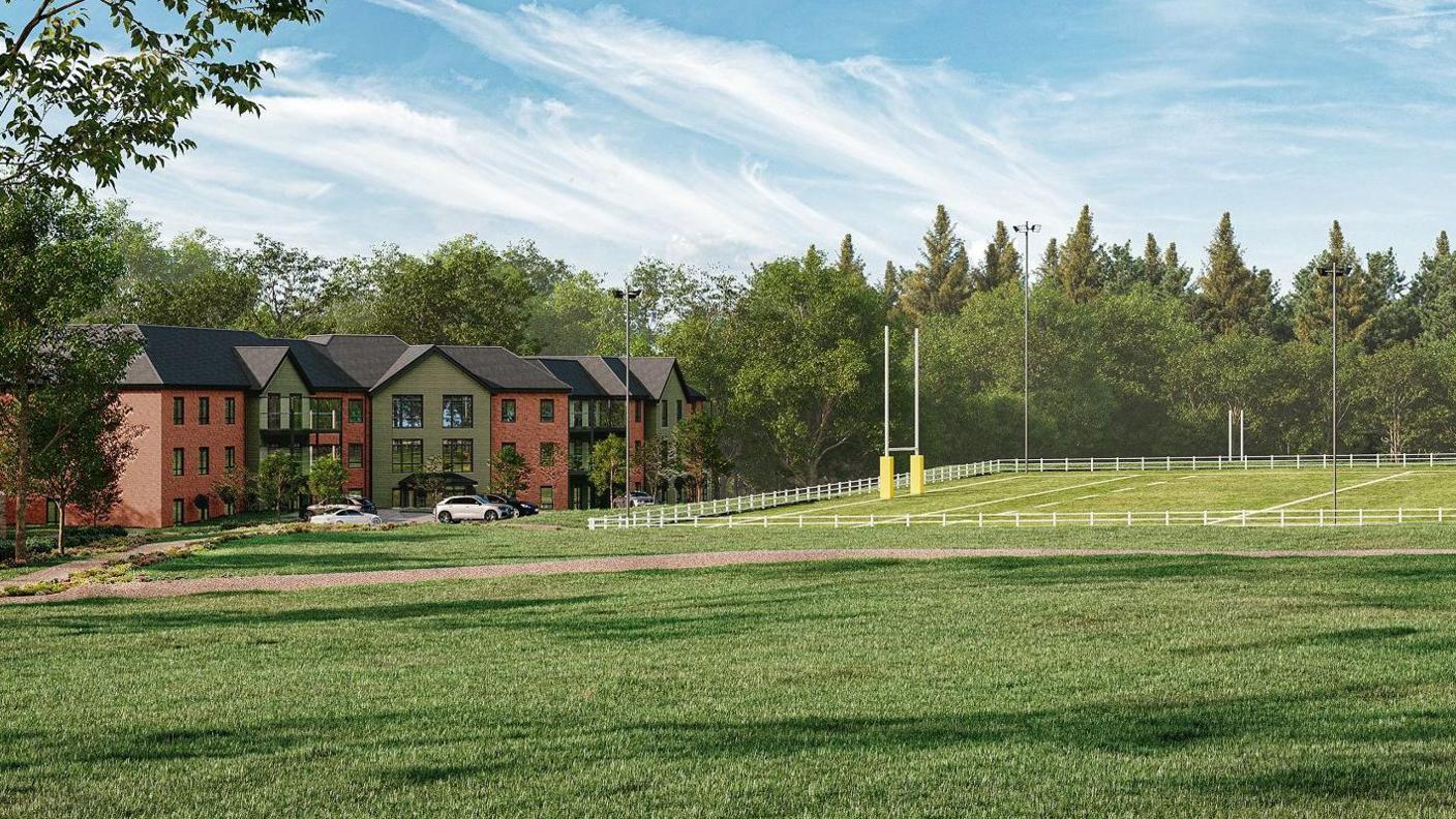 An architect's drawing of the proposed care home which has an alternate red brick and green wood facade, and is three storeys high with a grey roof. There are a few cars parked outside and it is right next to a rugby pitch. There is a rugby goal at one end and trees behind the pitch and care home, and a grass field in the foreground.