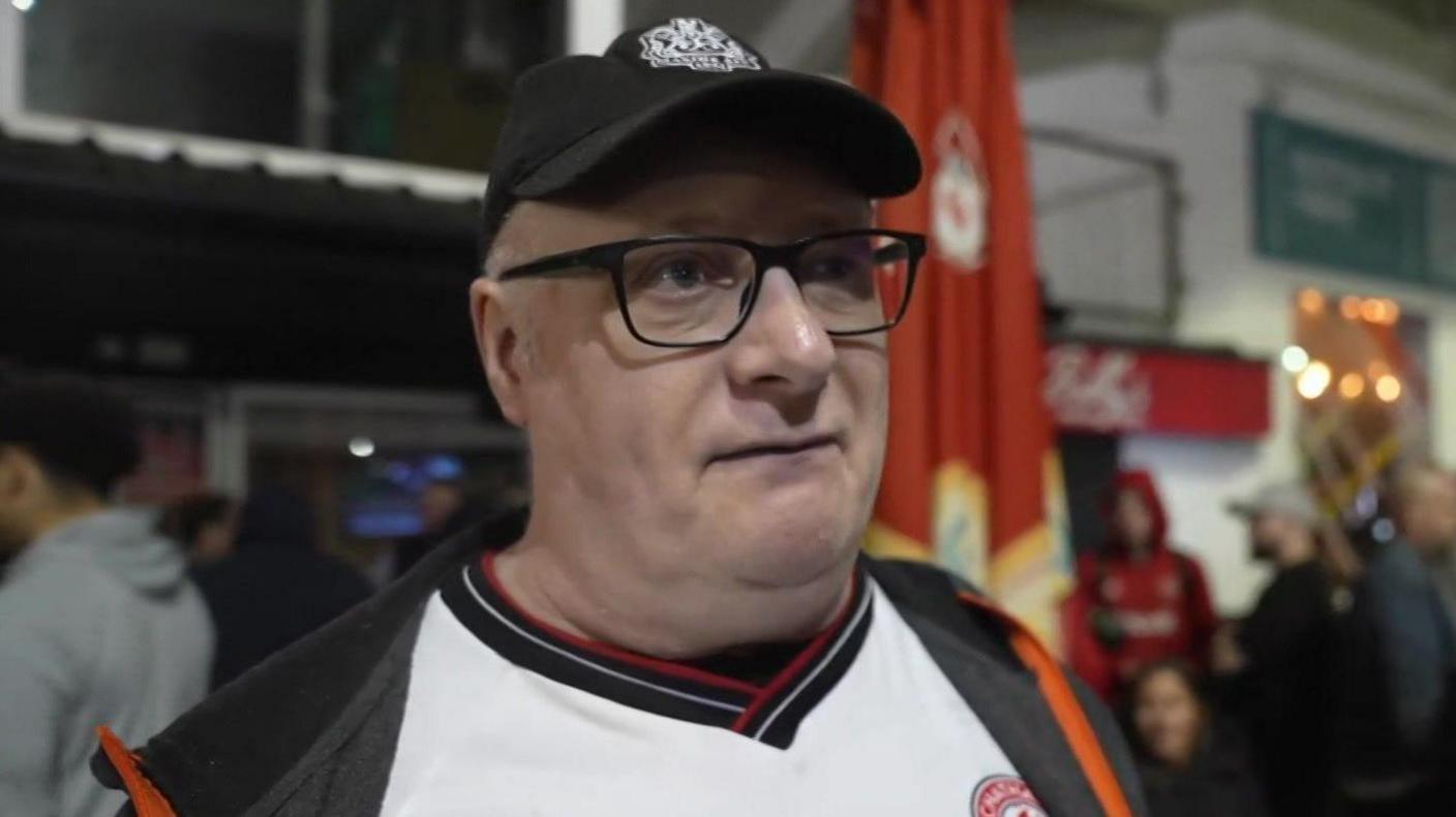 A man with black glasses and a black hat on. He is wearing a white Chatham Town shirt.