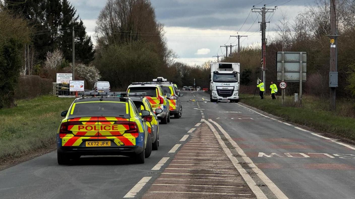 A row of police vehicles along the A1079 at Wilberfoss, with officers investigating at the scene. A white HGV is stationary on one side of the road. Two police officers with equipment in their hands are by its side.