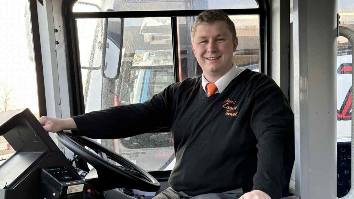 Dan Trent sits in the driver's cab of a coach and smiles at the camera. He has one hand resting on the steering wheel. He wears a black jumper with a white shirt and tie underneath. 