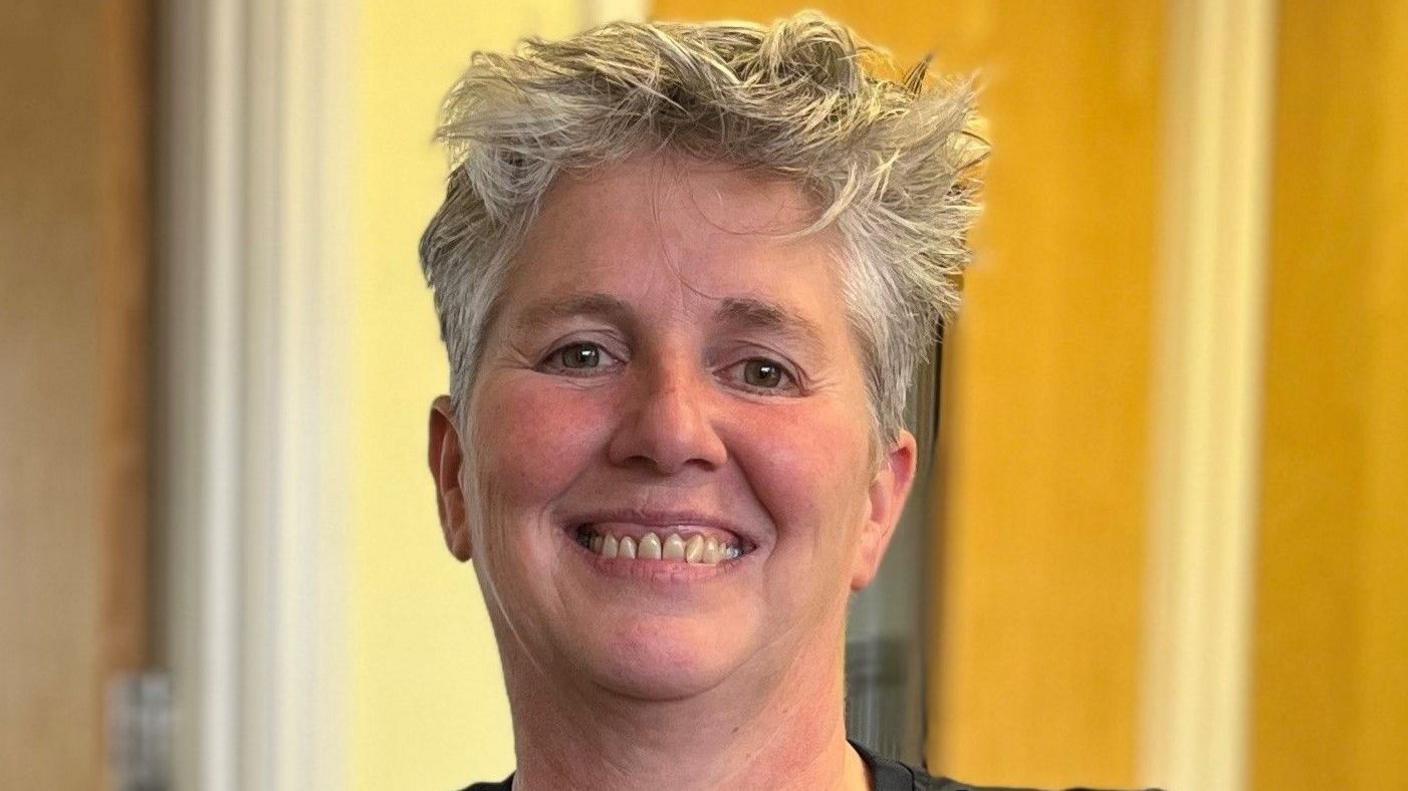 EJ, who has short grey hair, is standing in a room and smiling into the camera. Wooden doors are in the background.