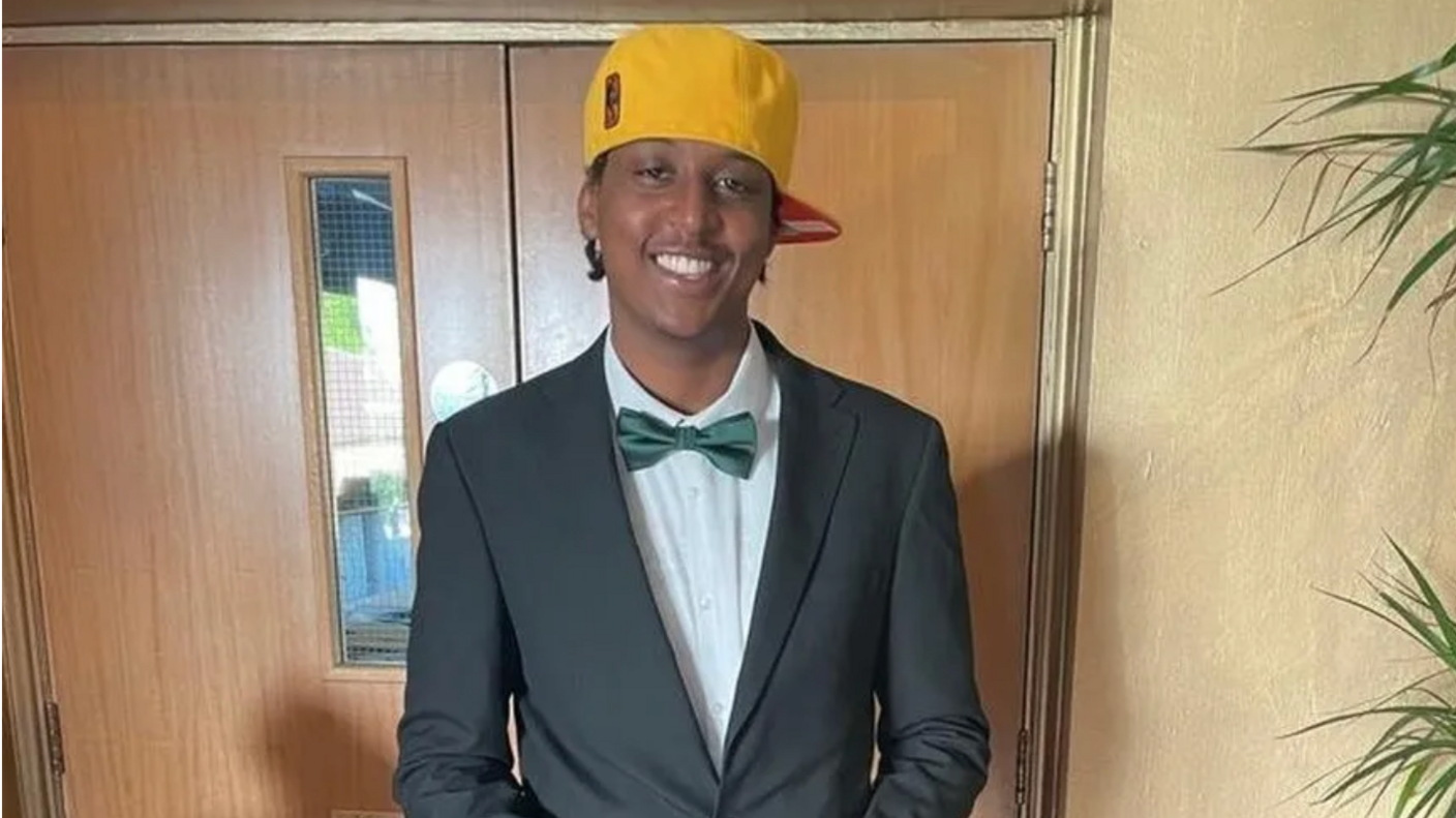 Eddie Kinuthia is standing in front of brown wooden swing doors. He is wearing a yellow baseball cap, a grey suit and green bow tie 