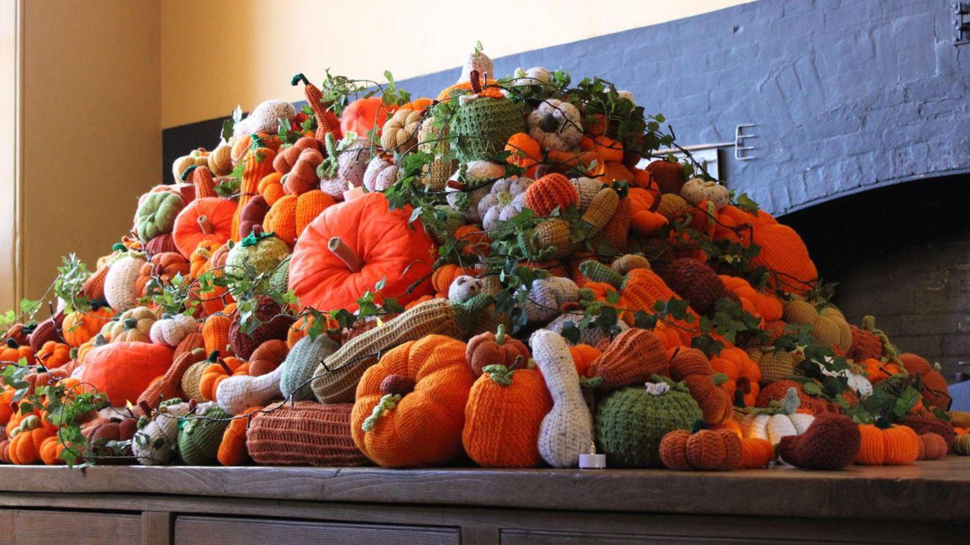 Hundreds of brightly coloured knitted pumpkins and seasonal autumnal vegetables piled up on a tabletop in a kitchen.