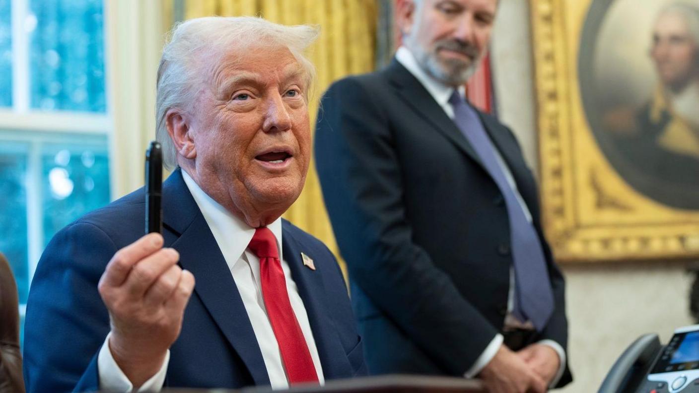 Donald Trump sitting behind a desk in the Oval Office, holding a pen aloft