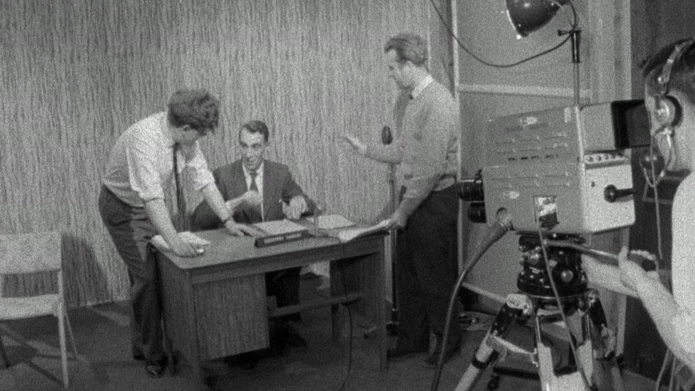 The newsreader Geoffrey Harvey, wearing a suit and tie, sitting behind a desk in the first Look East studio. He is speaking to two other men standing nearby. A camera and operator can be seen in the foreground.