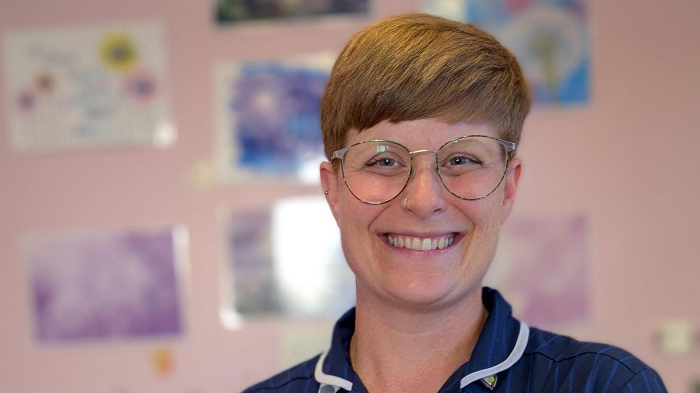 Sarah Cridland has short light brown hair and is smiling while wearing glasses and her blue midwife uniform.