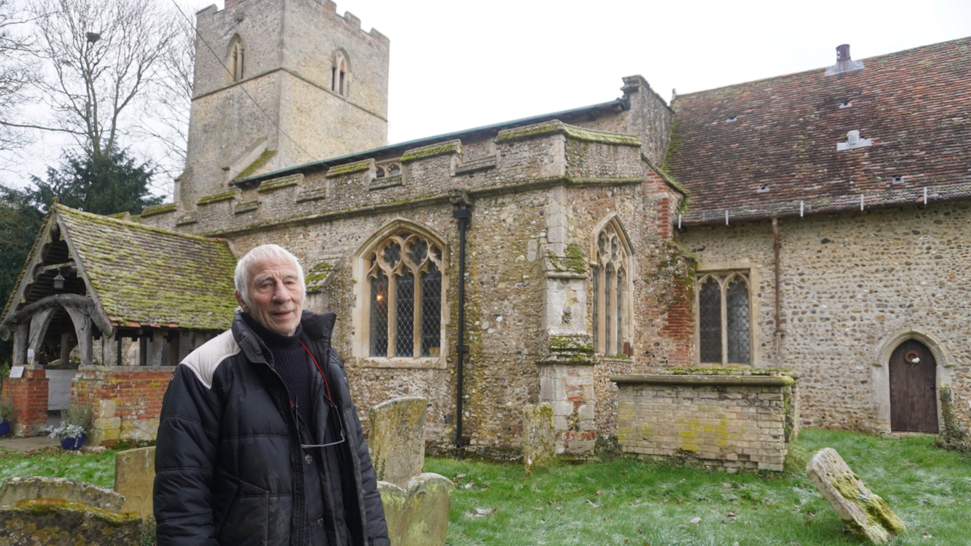 Churchwarden Peter Thompson, wearing a dark jumper and jacket, stands in the grounds of St Margaret's Church in Stradishall, with the entrance, side of the church and tower visible behind him.